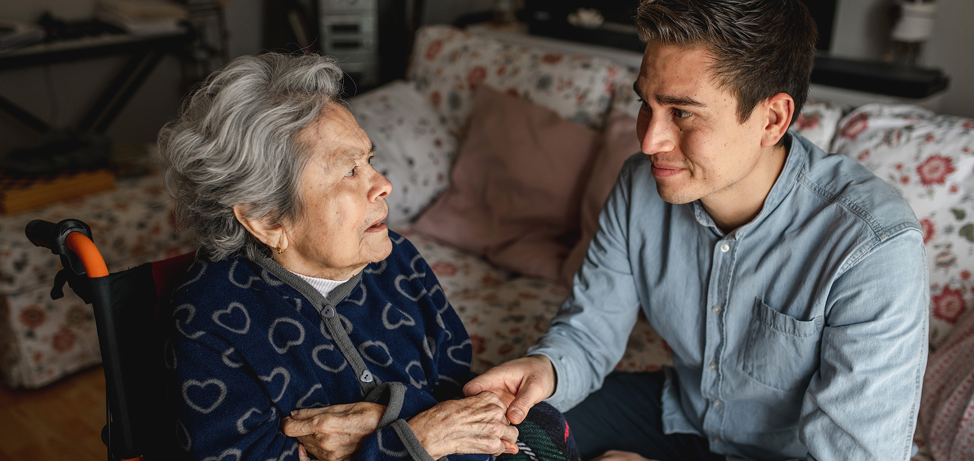 Older woman in wheel chair holding the hand of a younger man sitting on a couch