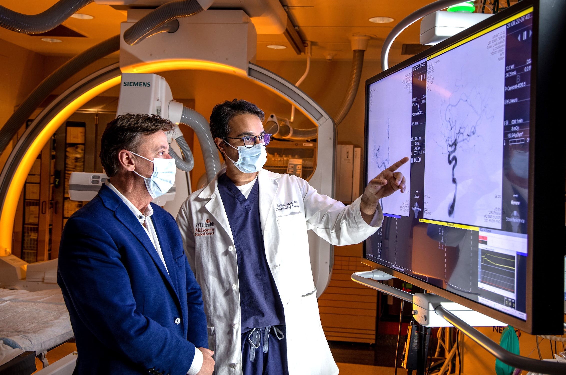 Two male doctors standing side-by-side wearing facemasks in a room of medical equipment, both looking at a medical monitor.