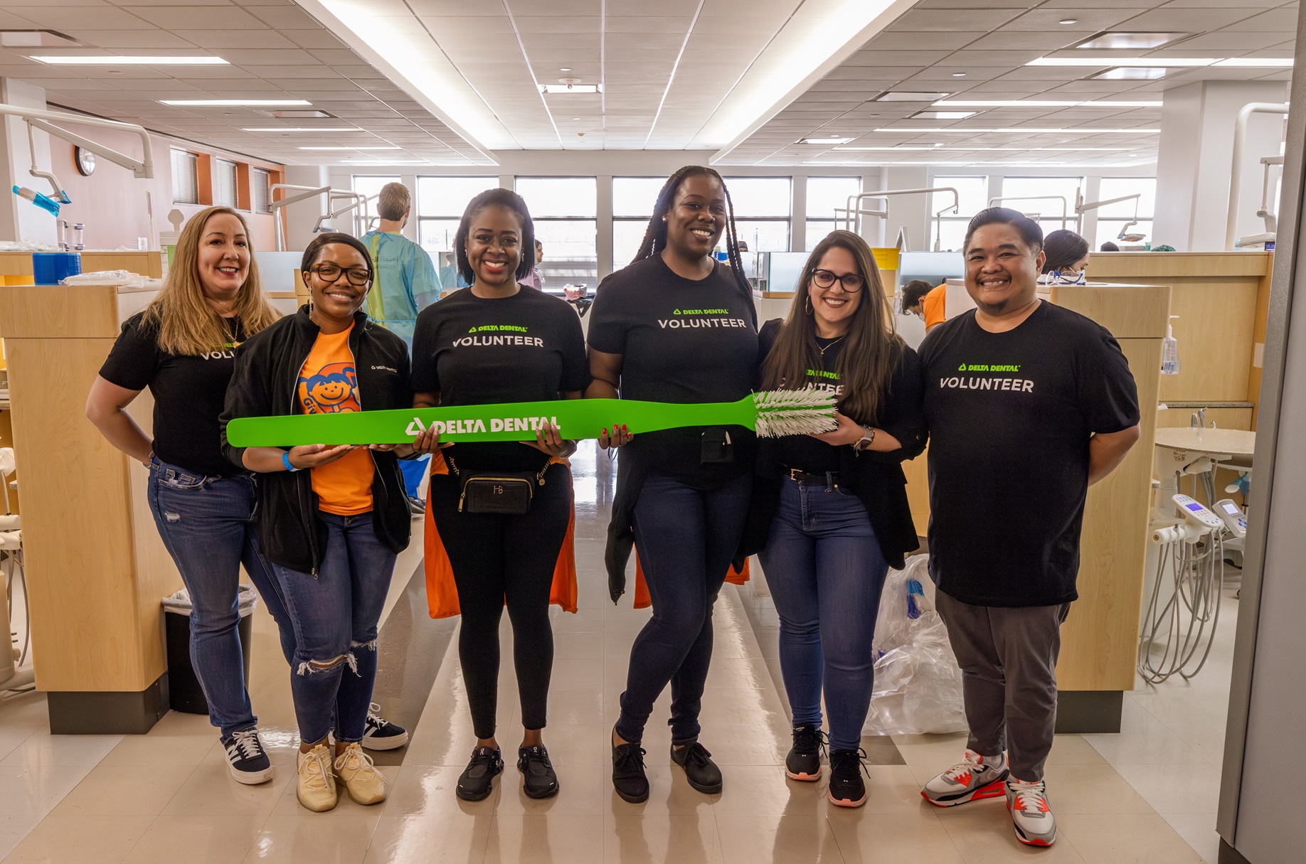 Group of six individuals standing side-by-side in a dental office with center four people holding a large, bright green, prop toothbrush with 'Delta Dental' written on the handle.