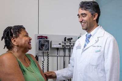 Aanand Naik, MD, who is the Executive Director for the Institute on Aging is standing and talking to a seated female patient on an examiniation table.