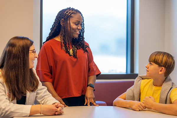 Candid photo of 3 individuals around a large table. At the far left is a female doctor (seated) and beside her is a young woman (standing.) Seated on the far right is a young boy. They all appear to be caught in a moment of conversation.